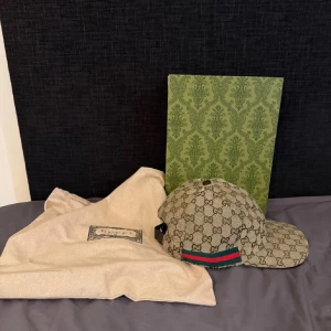 Gucci keps med monogram och gröna/röda detaljer - Säljer en klassisk Gucci keps med beige och brunt GG-monogram, gröna och röda ränder på sidan samt justerbart spänne bak i brunt läder. Kepsen är i canvasmaterial och kommer med dustbag och box. 