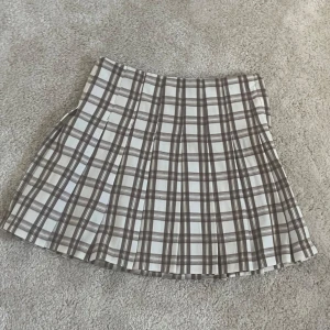 Brun och vit rutig kortkjol från H&M - Superfin kortkjol från H&M i brun och vit rutig design. Kjolen har dragkedja i sidan och är lätt utställd med plisserade veck. Perfekt för en trendig och stilren look!