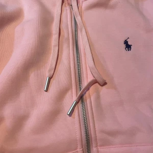 Ljusrosa hoodie från Polo Ralph Lauren - Ljusrosa hoodie från Polo Ralph Lauren med dragkedja och justerbar huva. Klassisk broderad logga på bröstet och silverfärgade metalldetaljer på snörena. Perfekt för en avslappnad och stilren look.