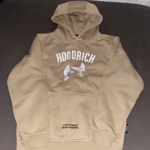 Beige väst från Hoodrich med tryck - Säljer en beige väst från Hoodrich med vita duvor och texttryck både fram och bak. Hoodien har en huva och känguruficka framtill samt flera snygga detaljer med svart text. Perfekt för en streetwear-look.
