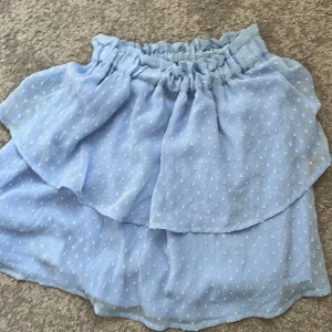 Ljusblå volangkjol från H&M - Supersöt ljusblå kjol från H&M med volangdetaljer och små diskreta prickar i tyget. Kjolen har resår i midjan för en bekväm passform och är tillverkad i ett lätt och luftigt material. Perfekt för varma dagar!