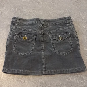 Grå manchesterkjol med fickor - Säljer en grå kort manchesterkjol med två fickor bak med knappar och klassisk jeansknäppning fram. Kjolen har bälteshällor och en rak passform. Perfekt för dig som gillar en enkel men snygg stil.