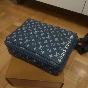 Blå axelväska från Louis Vuitton - Säljer en snygg blå axelväska från Louis Vuitton med det klassiska monogrammönstret i vitt. Väskan har en praktisk dragkedja och är perfekt för att bära dina dagliga nödvändigheter. En stilren accessoar för alla tillfällen.