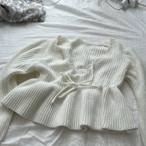 Vit stickad tröja med knyt - Säljer en söt vit stickad tröja med knyt i midjan och volangdetaljer vid halsen. Tröjan har långa ärmar och en ribbad struktur som ger en fin passform. Perfekt för kyliga dagar!