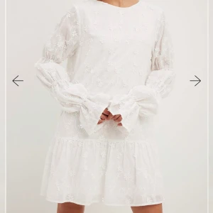 Vit blommig klänning med öppen rygg - En vit klänning med blommigt mönster och öppen rygg. Den har långa ärmar med volangdetaljer och en knytning i ryggen. Perfekt för en somrig look!