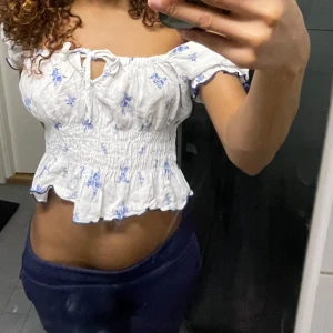 Vit blommig offshoulder topp - Söt vit offshoulder topp med blå blommor och volangdetaljer. Toppen är croppad och har en smockad midja för en bekväm passform. Perfekt för varma dagar!