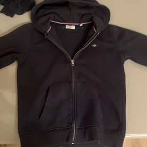 Mörkblå hoodie från GANT - Säljer en snygg mörkblå hoodie från GANT med dragkedja och huva. Den har en liten logga på bröstet. Perfekt för en casual look! 