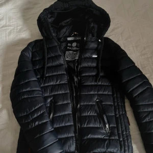 Marinblå/svart pufferjacka från Navahoo - Säljer en svart pufferjacka från Navahoo med huva och dragkedja. Jackan har en quiltad design och praktiska fickor med dragkedjor. Perfekt för kyliga dagar.