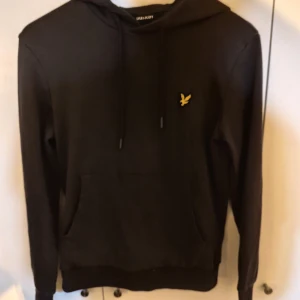 Svart hoodie från Lyle & Scott - Säljer en stilren svart hoodie från Lyle & Scott med deras ikoniska logga på bröstet. Tröjan har en klassisk design med dragsko i huvan och en praktisk magficka. Perfekt för en avslappnad look. Står xs men passar s