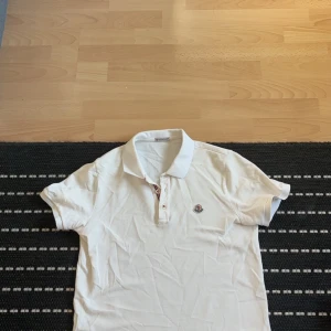 Vit kortärmad pikétröja från Moncler - Säljer en stilren vit pikétröja från Moncler med korta ärmar och klassisk krage. Tröjan har en broderad logotyp på bröstet och knappar vid halsen. Perfekt för en avslappnad men ändå snygg look.