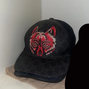 Gucci keps - Snygg svart keps med en iögonfallande röd vargdesign framtill. Kepsen har ett diskret mönster i bakgrunden och justerbar passform. Perfekt för att ge din outfit en cool touch. Nästan omöjlig få tag på