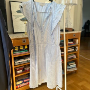 Blå- och vitrandig klänning från InWear - Säljer en somrig blå- och vitrandig klänning från InWear. Klänningen har en v-ringad design med knappar framtill och volangdetaljer vid ärmarna. Tillverkad i 100% bomull, perfekt för varma dagar. Den har en liten fläck därav lågt pris.