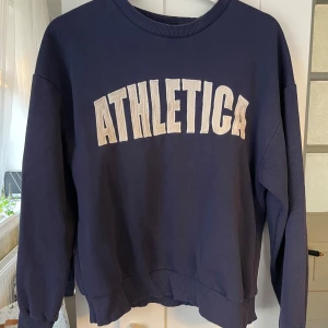 Mörkblå sweatshirt Gina Tricot - Säljer en mörkblå sweatshirt från Gina Tricot med texten 'ATHLETICA' i vitt på framsidan. Tröjan har långa ärmar och ribbade muddar vid hals, ärmslut och nederkant. Perfekt för en avslappnad stil.