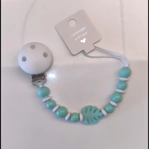 Handgjord napphållare med turkosa pärlor - Söt handgjord napphållare med turkosa och vita pärlor samt ett bladformat dekorativt element. Perfekt för att hålla nappen nära och säker. Klämman är vit och rund med hål för enkel fastsättning.