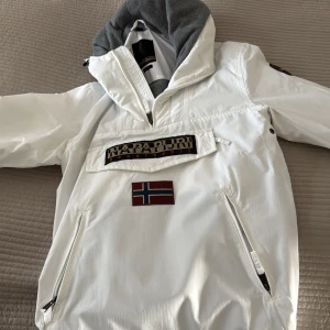 Vit anorak från Napapijri - Säljer en stilren vit anorak från Napapijri med en stor ficka framtill och en norsk flagga som detalj. Jackan har en justerbar huva och dragkedjor vid fickorna. Perfekt för kyligare dagar.