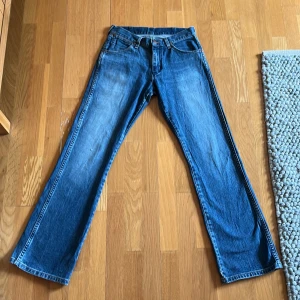 Blå jeans från Wrangler - Säljer ett par klassiska blå jeans från Wrangler. De har en bootcut-stil och är tillverkade i slitstarkt denim. Jeansens baksida har de ikoniska Wrangler-detaljerna med lädermärke och kontrastsömmar.