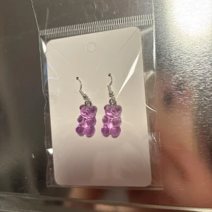 Lila örhängen med nallebjörnar - Söta örhängen med lila nallebjörnar i plast. Perfekta för att lägga till en lekfull touch till din outfit. De hänger från silverfärgade krokar och är lätta att bära. (Billigt material) finns 2 par. 