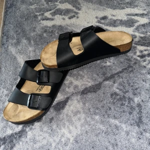 Svarta sandaler från Birkenstock - Säljer ett par klassiska svarta sandaler från Birkenstock med två justerbara remmar och en bekväm korksula. Perfekta för sommarens alla äventyr. Sandalerna har en ergonomisk fotbädd för extra komfort.