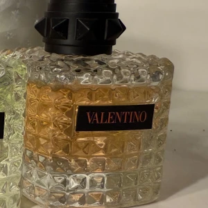 Parfym Corall - Elegant parfymflaska från Valentino med en lyxig design. Flaskan har en unik, strukturerad yta i glas med en svart etikett och texten 'VALENTINO'. Korken är svart med geometriska detaljer. Perfekt för den som vill ha en stilren och exklusiv doftupplevelse.