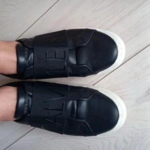 Svarta sneakers från Calvin Klein - Snygga svarta sneakers från Calvin Klein med breda elastiska band över foten och vit sula. Skorna har en stilren design med märkets logga både på bandet och hälen. De är använda men ser ändå hyfsatt nya ut. Strl 37 men passar mig som har strl 36. 