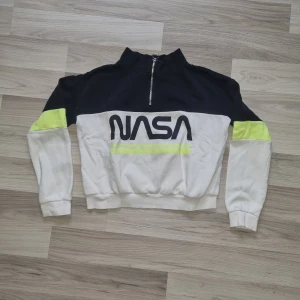 Svart och vit tröja med NASA-tryck från H&M - Cool tröja från H&M med NASA-tryck i svart och vit. Den har en dragkedja framtill och neongula detaljer på ärmarna. Perfekt för en avslappnad stil med en modern touch.