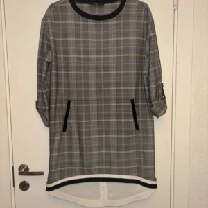 Rutig klänning från Say - Ej använd. Snygg rutig klänning från Say i grå och svart med trekvartsärm och rund hals. Klänningen har svarta detaljer vid fickorna och en kontrasterande kant vid fållen. Perfekt för en stilren look!
