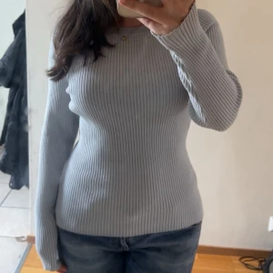 Ljusblå ribbad tröja från H&M - Säljer en tajt ljusblå ribbad tröja från H&M. Tröjan har långa ärmar och dekorativa knappar på axlarna. Perfekt för en elegant och bekväm look. 