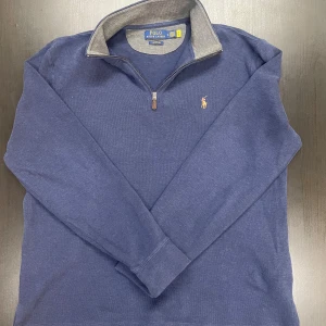 Blå tröja med dragkedja från Polo Ralph Lauren - Säljer en stilren blå tröja från Polo Ralph Lauren med dragkedja och broderad logga på bröstet. Tröjan har en klassisk krage och är perfekt för en avslappnad look. Passar M och är slim fit på L