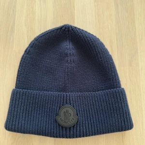 Moncler mössa mörkblå  - Snygg mörkblå mössa från Moncler med ribbad design och uppvikt kant. Perfekt för kyliga dagar med stilren look. Moncler-logga framtill ger en exklusiv touch. Mössan är aldrig använd och är helt ny som ni ser på bilderna, tagsen till mössan finns kvar och skickas med vid köp! 