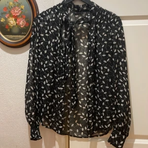Very pretty🖤🤍 - "This black sheer blouse with the white floral print is absolutely stunning! The long sleeves with elastic cuffs add a nice touch, and I love how it hangs so elegantly in front of that white door. It really creates a beautiful contrast. Do you think this would be perfect for a night out, or maybe a more casual day look🖤🤍