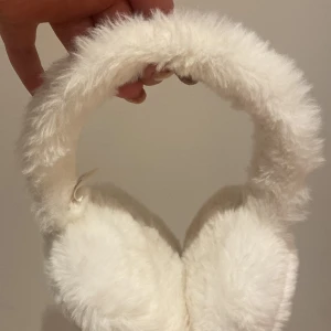 Vita pälsiga öronmuffar från Jyothi - Säljer ett par supermysiga vita öronmuffar från Jyothi. De är täckta med mjuk päls och perfekta för att hålla värmen under kalla dagar. En stilren accessoar för vintern!