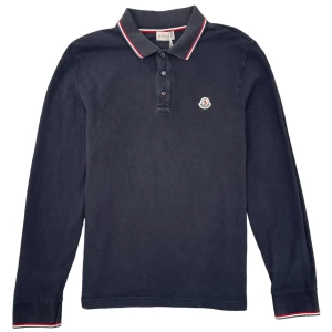 Svart pikétröja från Moncler - Snygg svart pikétröja från Moncler med långa ärmar och klassisk krage. Tröjan har en liten logotyp på bröstet och röd-vit randdetalj vid krage och ärmslut. Perfekt för en stilren look.