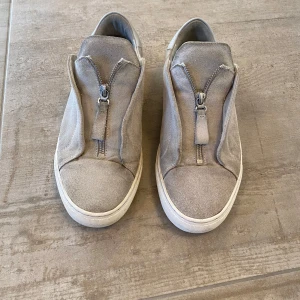 Grå sneakers med dragkedja - Snygga grå sneakers i mocka med dragkedja framtill. De har en rund tå och en vit sula som ger en stilren look. Perfekta för en avslappnad stil. Skriv om ni har frågor om skorna.