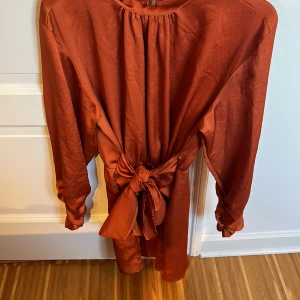Rostfärgad klänning med knytband - Snygg rostfärgad klänning med knytband i midjan och långa ärmar. Perfekt för höst och vinter med sin varma ton. Klänningen har en enkel design med en liten öppning i nacken.