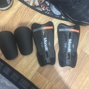 Svarta benskydd från Monolit - Säljer ett par svarta benskydd från Monolit. De är 14 cm långa och har en slimmad design med en hård yta för extra skydd. Perfekta för fotbollsträning eller match.