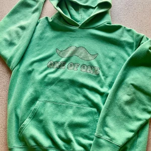 Grön hoodie med text - Säljer en grön hoodie med en unik design. Den har en mustasch och texten 'ONE OF ONE' på framsidan. Perfekt för en avslappnad stil. Perfekt tull sommaren till sommaren. Hoodien är mycket nopprig därför billigt pris.