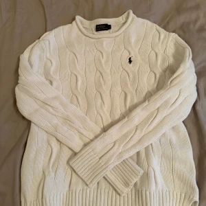 Vit stickad tröja från Polo Ralph Lauren - Säljer en stilren vit stickad tröja från Polo Ralph Lauren med klassiskt flätmönster. Tröjan har långa ärmar och en broderad logotyp på bröstet. Perfket och skön för Sverige väder!