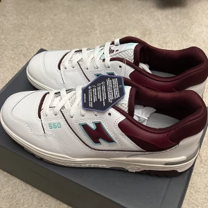 New Balance 550 i vitt och vinrött  - Snygga New Balance 550 sneakers i vitt med vinröda detaljer. Skorna har en klassisk design med snörning och en bekväm passform. Perfekta för både vardag och träning.