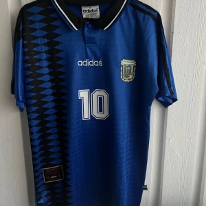 Blå fotbollströja från Adidas - Säljer en blå fotbollströja från Adidas med nummer 10 och texten 'Maradona' på ryggen. Tröjan har ett mönster av svarta och blå trianglar på sidan och korta ärmar. Perfekt för fotbollsfans!
