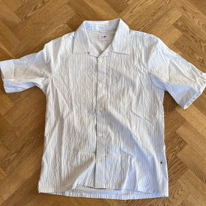 Ljusblå randig skjorta från NN07 - Säljer en stilren ljusblå skjorta från NN07 med smala vita ränder. Skjortan har korta ärmar och knappar framtill. Perfekt för en avslappnad men ändå snygg look. 🕶️