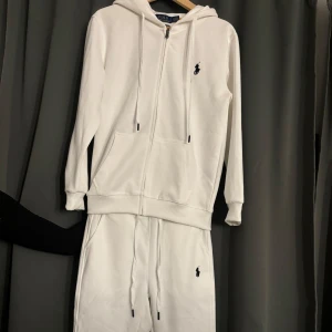 Vit hoodie från Ralph Lauren - Säljer en stilren vit tracksuit med dragkedja och det klassiska logotypbroderiet på bröstet. Tröjan har långa ärmar och en praktisk huva med snörning. Perfekt för en avslappnad look. Endast prövat 1 gång! 