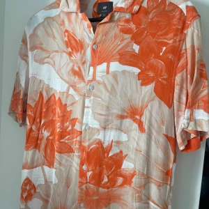 Orange blommig skjorta från H&M - Säljer en somrig kortärmad skjorta från H&M med ett stort blommönster i orange och beige. Skjortan har knappar framtill och en klassisk krage. Perfekt för en utekväll inför sommar/vår, stylea med linnébyxor för en elegant stil. Nypris: 499kr