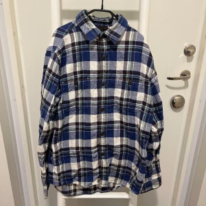 Blå och vit rutig skjorta - Säljer en snygg blå och vit rutig flanellskjorta med knappar framtill. Skjortan har en klassisk krage och långa ärmar. Storlek M