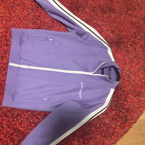Lila track tröja från Palm Angels - Snygg lila tröja från Palm Angels med vita ränder längs ärmarna och dragkedja framtill. Tröjan har en broderad logga på bröstet och är tillverkad i ett mjukt syntetmaterial. Ganska bra skick 