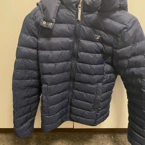 Blå dunjacka från Gant - Säljer en snygg blå dunjacka från Gant med dragkedja och huva. Jackan har en quiltad design och är perfekt för kyligare väder. Den har en stilren look och är både praktisk och trendig.