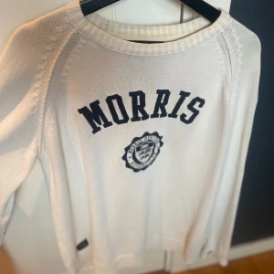 Vit tröja från Morris - Säljer en stilren vit tröja från Morris med mörkblå text och emblem på framsidan. Tröjan har långa ärmar och mörkblå armbågslappar som ger en snygg kontrast. 