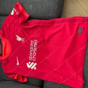 Liverpool tröja från Nike - Säljer en röd fotbollströja från Nike med vita detaljer och klubbmärke på bröstet. Tröjan har korta ärmar och ett stilrent mönster. Perfekt för fotbollsfans!