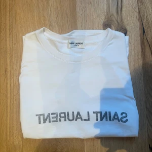 Vit t-shirt från Saint Laurent - Säljer en stilren vit t-shirt från Saint Laurent med deras ikoniska logga tryckt på framsidan. Perfekt för en minimalistisk look. T-shirten har en klassisk passform och är kortärmad.