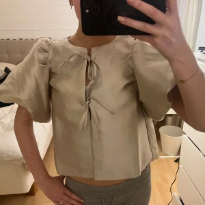 Blus med puffärmar - Säljer en snygg champagne färgad blus från Gina Tricot i storlek Xs! Den har puffiga korta ärmar och knytning framtill. Bra skick, använd en gång. Blusen har en lös passform och är tillverkad i ett mjukt material.