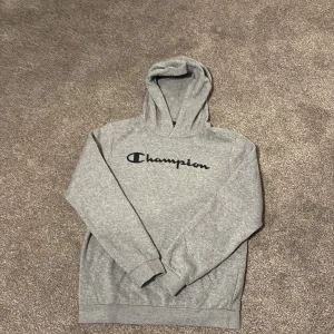 Grå hoodie från Champion - Säljer en klassisk grå hoodie från Champion med deras ikoniska logga tryckt på bröstet. Tröjan har en bekväm passform med långa ärmar och en praktisk huva. Perfekt för en avslappnad stil.